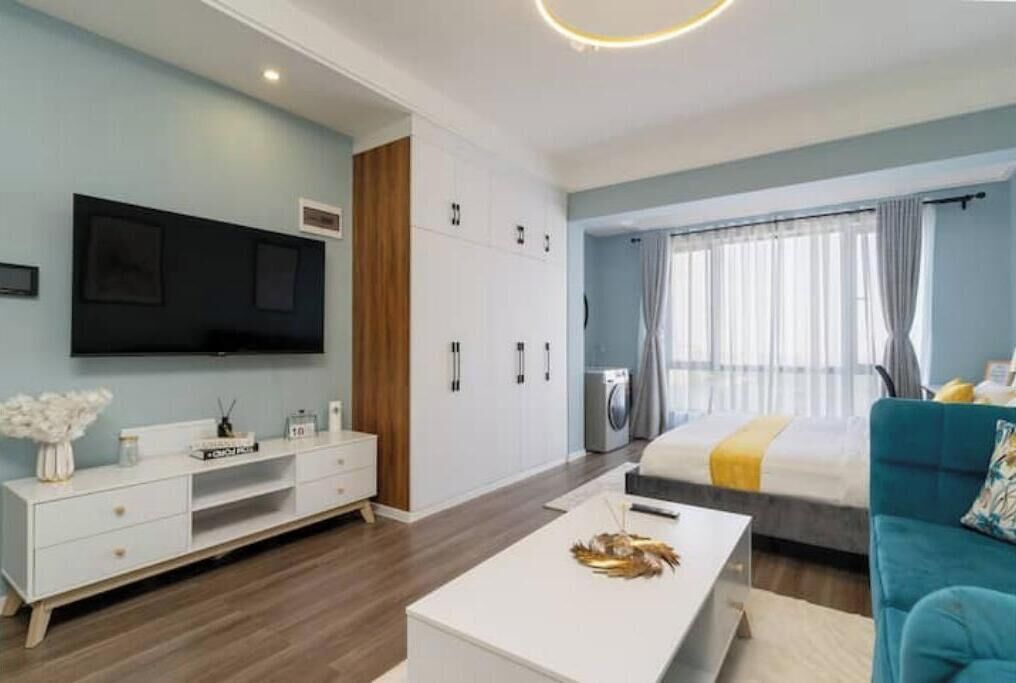 Ein modernes Hotelzimmer mit einem Flachbildfernseher, einem weißen Konsolentisch, einem Bett mit gelber Tagesdecke und einem blauen Sofa.