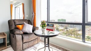 A modern living room with a brown leather sofa, a glass coffee table, a potted plant, and a city view through large windows. - Deluxe Haven Kilimani with pool & gym yaya center (Nairobi)