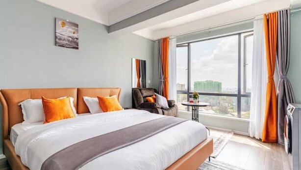 A modern hotel room with a large bed, a view of the city, and a seating area with a table and chairs. - Deluxe Haven Kilimani with pool & gym yaya center (Nairobi)