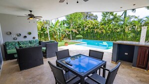 Une terrasse extérieure avec une table à plateau de verre et des chaises en osier, une piscine et une végétation luxuriante en arrière-plan.