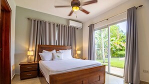 Une chambre à coucher avec un lit en bois, deux tables de chevet, un ventilateur de plafond et une grande fenêtre avec des rideaux.