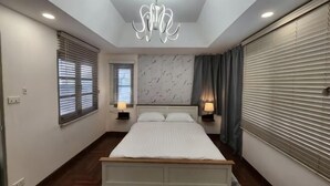 A bedroom with a bed, two bedside tables, a chandelier, and a window with blinds.