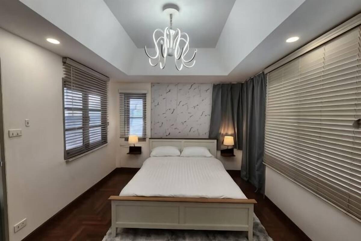 A bedroom with a bed, two bedside tables, a chandelier, and a window with blinds.
