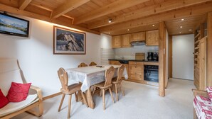 Ein Holzinnenraum mit Esstisch, Stühlen, einer Küchenzeile und einem Bett mit rotem Kopfkissen.