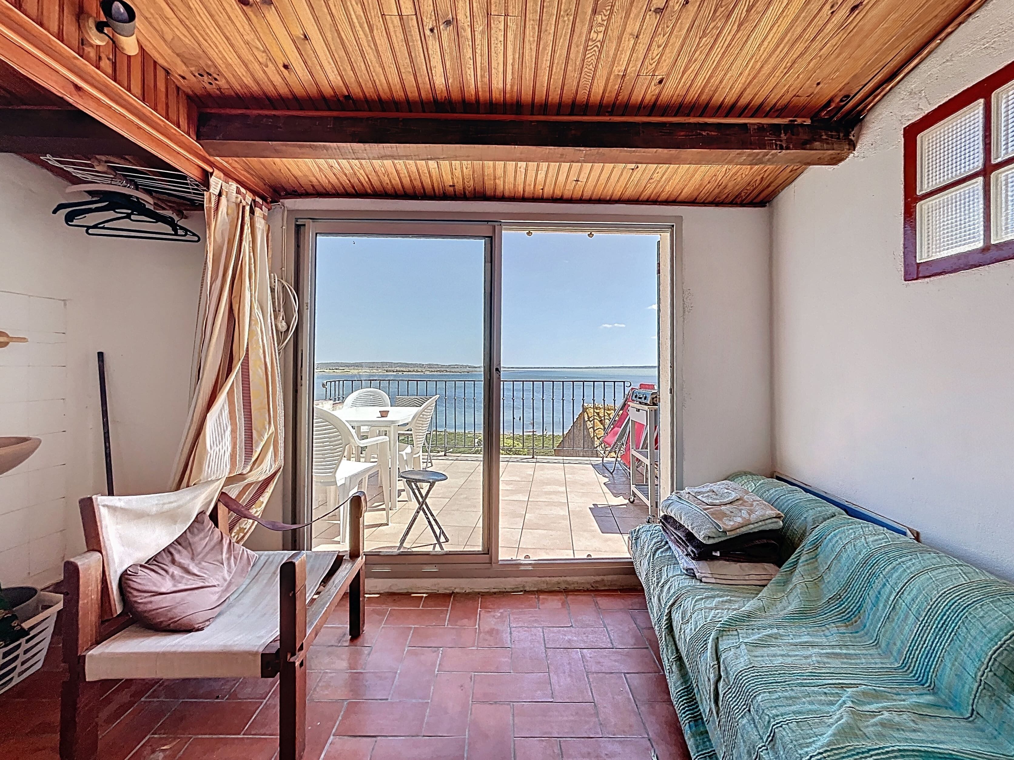 A room with a wooden ceiling, a sliding glass door leading to a balcony with a table and chairs, and a bed with a patterned cover.