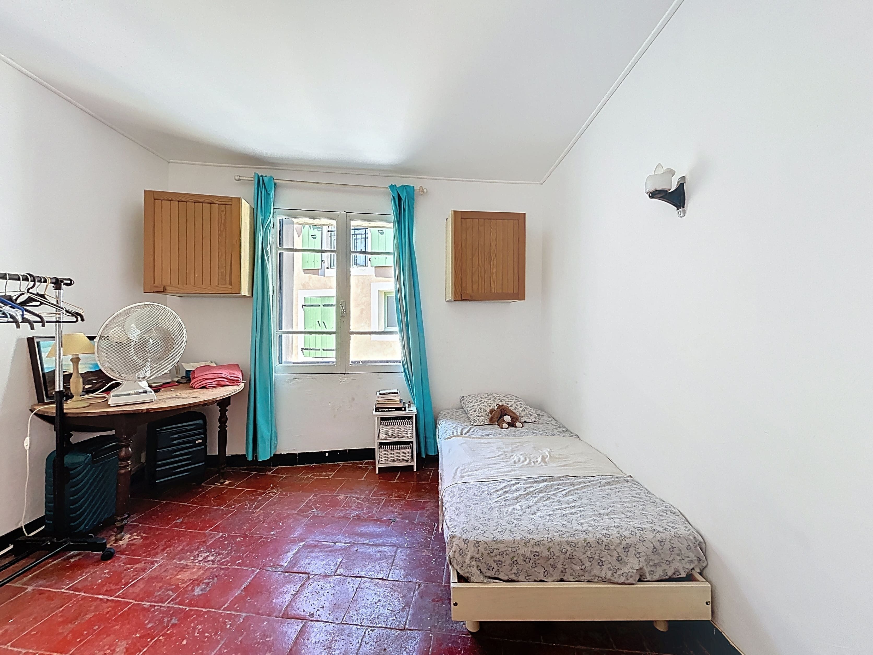 Een kleine, compacte kamer met een bed, een bureau met een ventilator, een klein tafeltje en een raam met gordijnen.