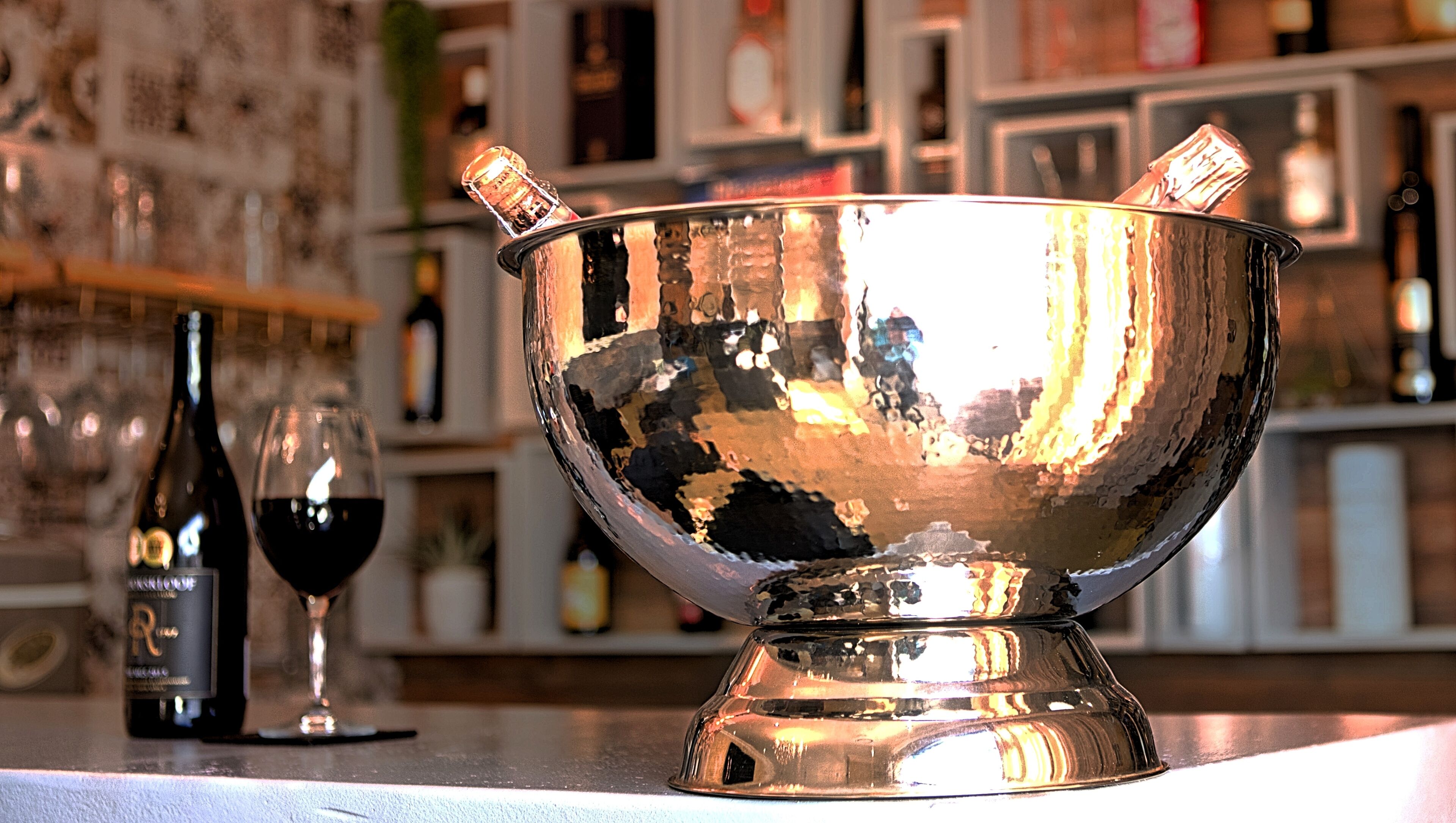Un bar con un tazón metálico reflectante, dos copas de vino y una botella de vino tinto.