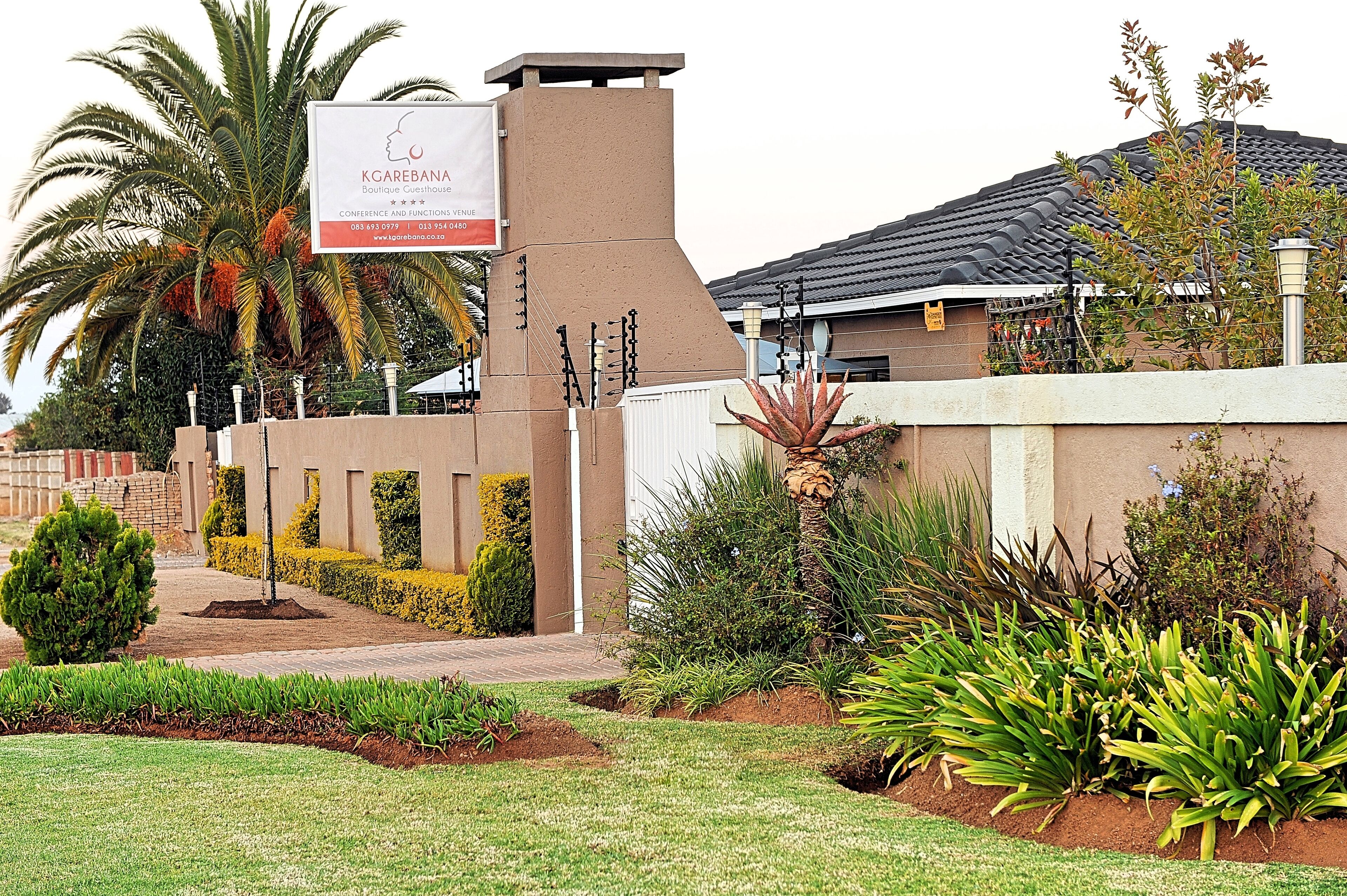 A well-maintained garden with palm trees and various shrubs, a signboard for 'Kagebana Boutique Guesthouse', and a modern building with a tiled roof.