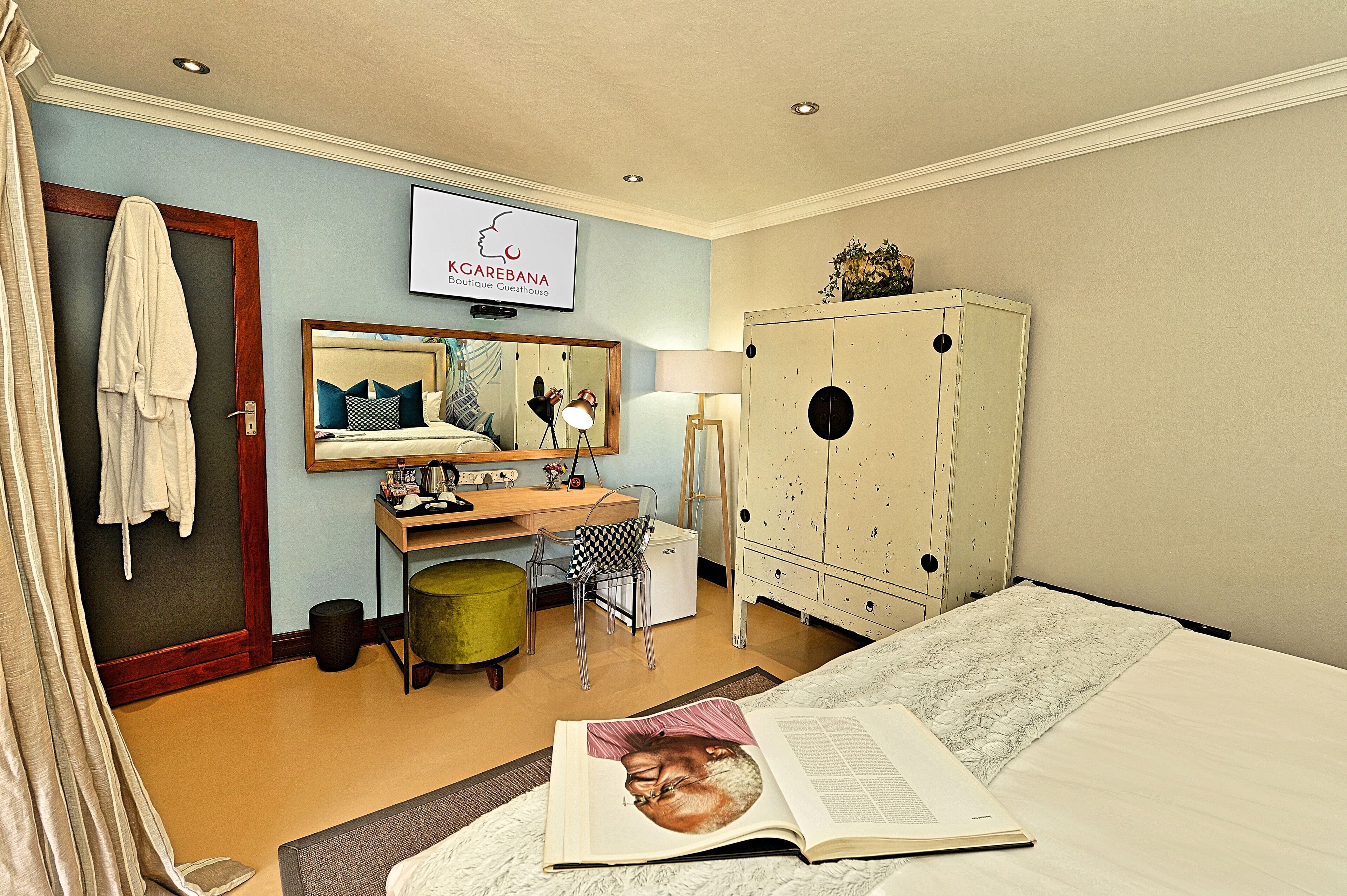 A hotel room with a bed, a desk with a chair, a wardrobe, a mirror, and a TV displaying a sign.