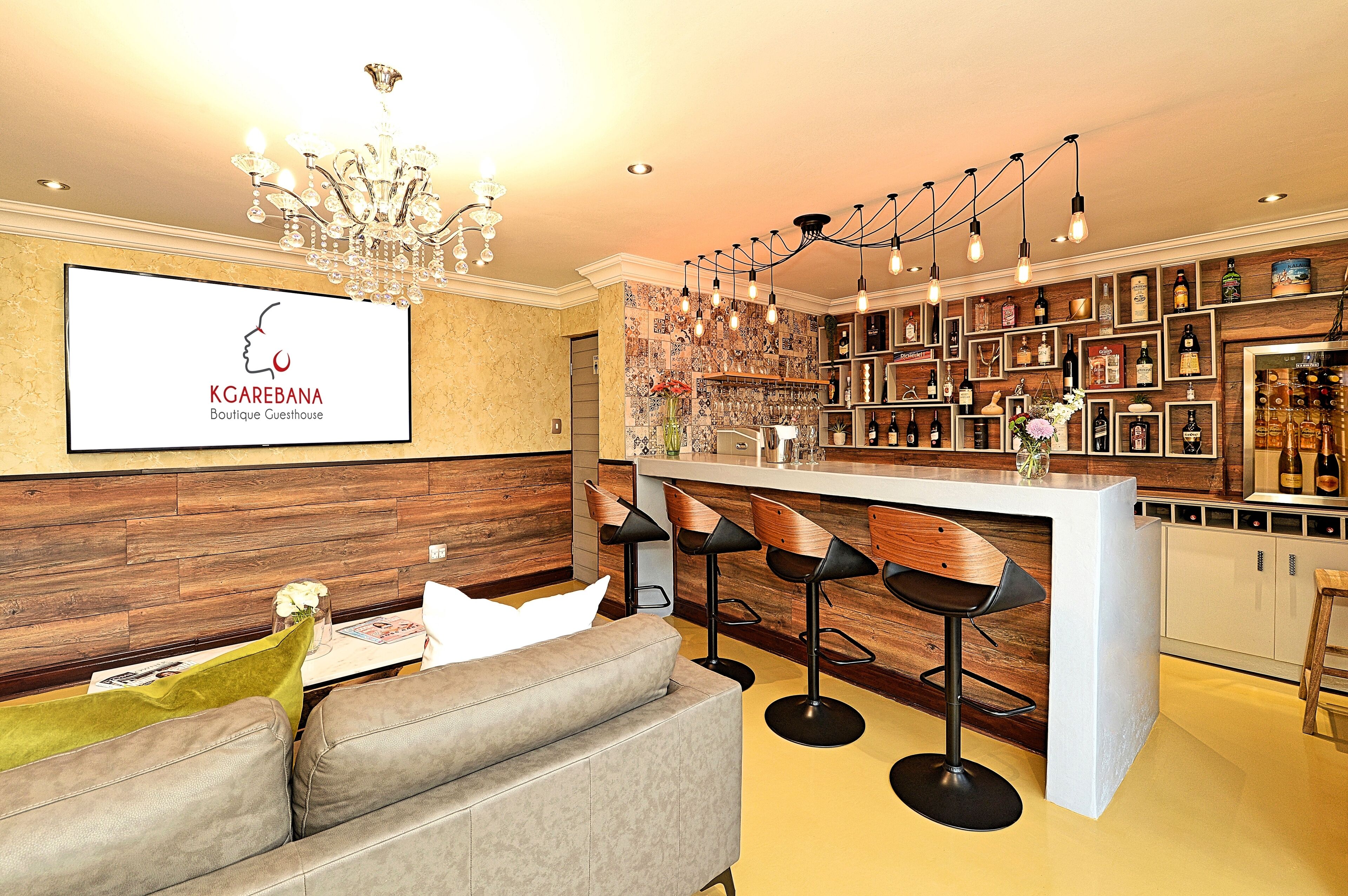 A modern bar area with a chandelier, bar stools, and a variety of bottles and glasses on shelves.
