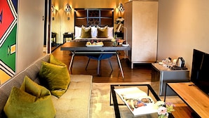 A modern hotel room with a wooden dining table, a sofa with green cushions, a flat-screen TV, and a minibar. - Kgarebana Boutique Guesthouse (Sesakhile)