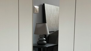 Una habitación con un armario blanco, una mesita de noche negra con una lámpara y una cama con cabecero gris.