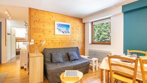 A cozy room with a wooden wall, a grey sofa, a small table with a chessboard, and a dining area with wooden chairs and a table. - OBIOU - 200m des Pistes de Ski (Chamrousse)