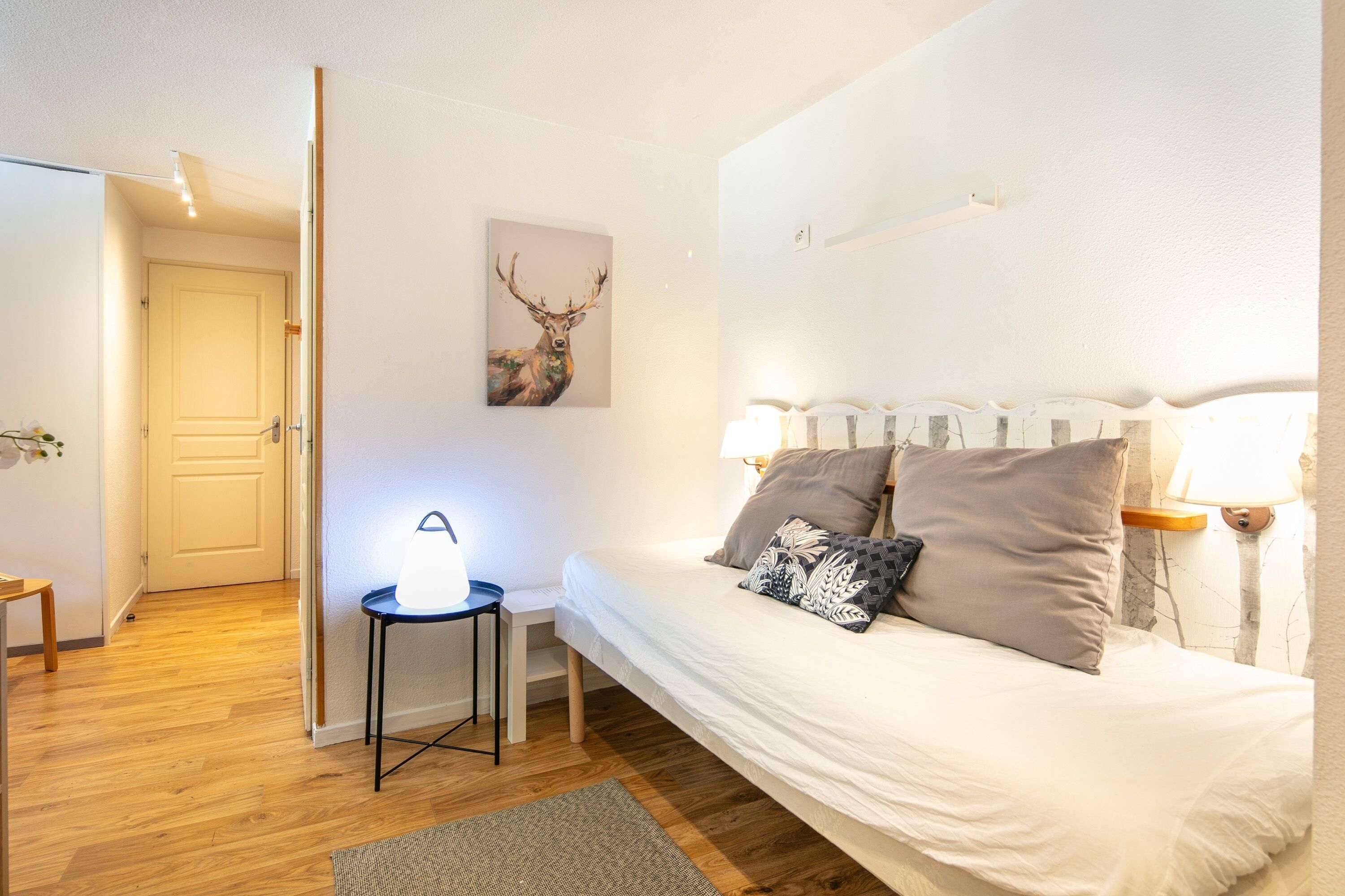 Un dormitorio con cama, lámparas de noche, una mesita con lámpara, un cuadro en la pared con un ciervo, y una puerta que da a otra habitación.