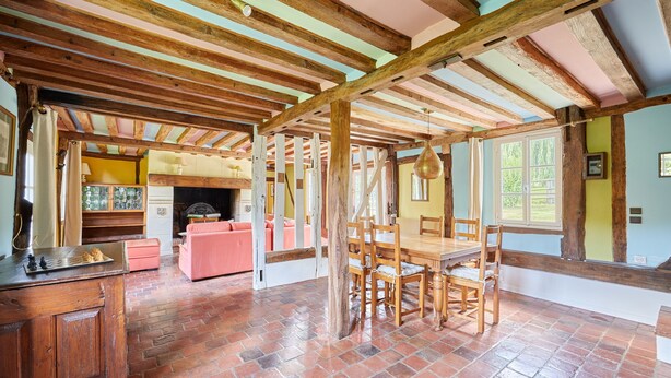 A rustic interior with exposed wooden beams, a brick floor, a dining area with wooden chairs, and a fireplace.