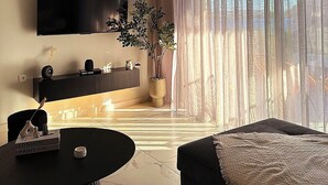 Ein modernes Wohnzimmer mit Flachbildfernseher, rundem Couchtisch, einer Couch, einer Topfpflanze und einem großen Fenster mit leichtem Vorhang.