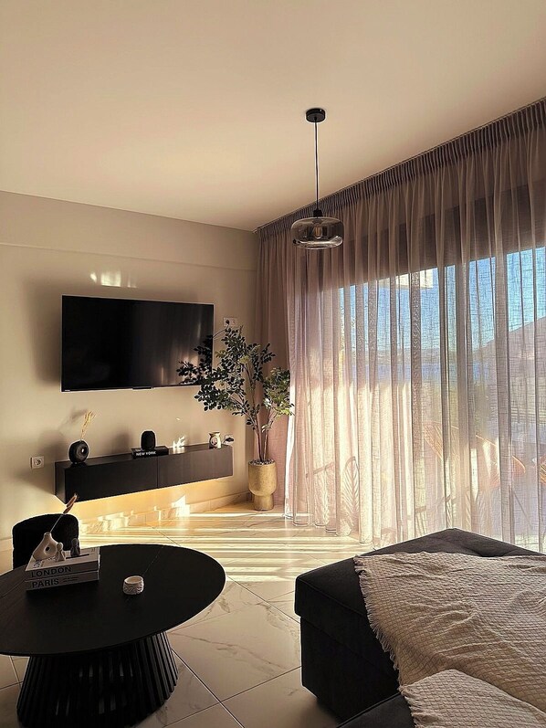 A modern living room with a flat-screen TV, a round coffee table, a sofa, a potted plant, and a large window with sheer curtains.
