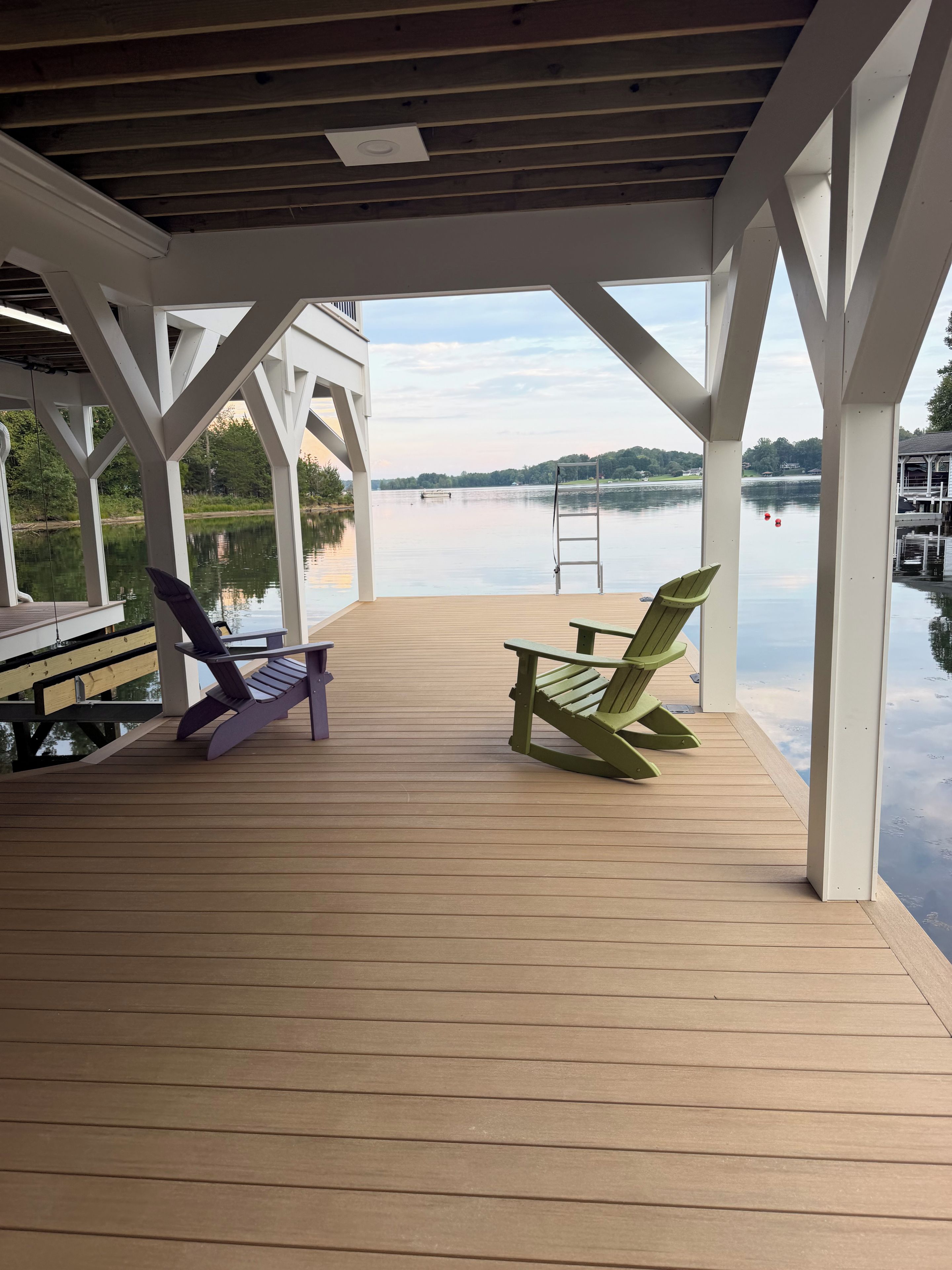 Une terrasse en bois couverte, avec deux fauteuils Adirondack, une échelle et une vue sur un lac paisible.