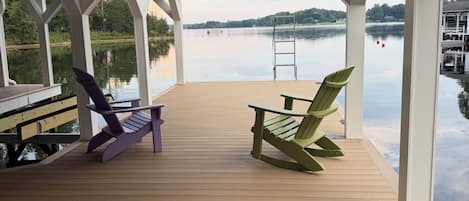 A covered wooden deck with two Adirondack chairs, a ladder, and a view of a calm lake.