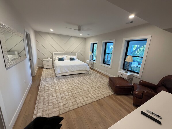 Un dormitorio moderno con una cama grande, mesitas de noche, un ventilador de techo y vistas al exterior a través de ventanas.