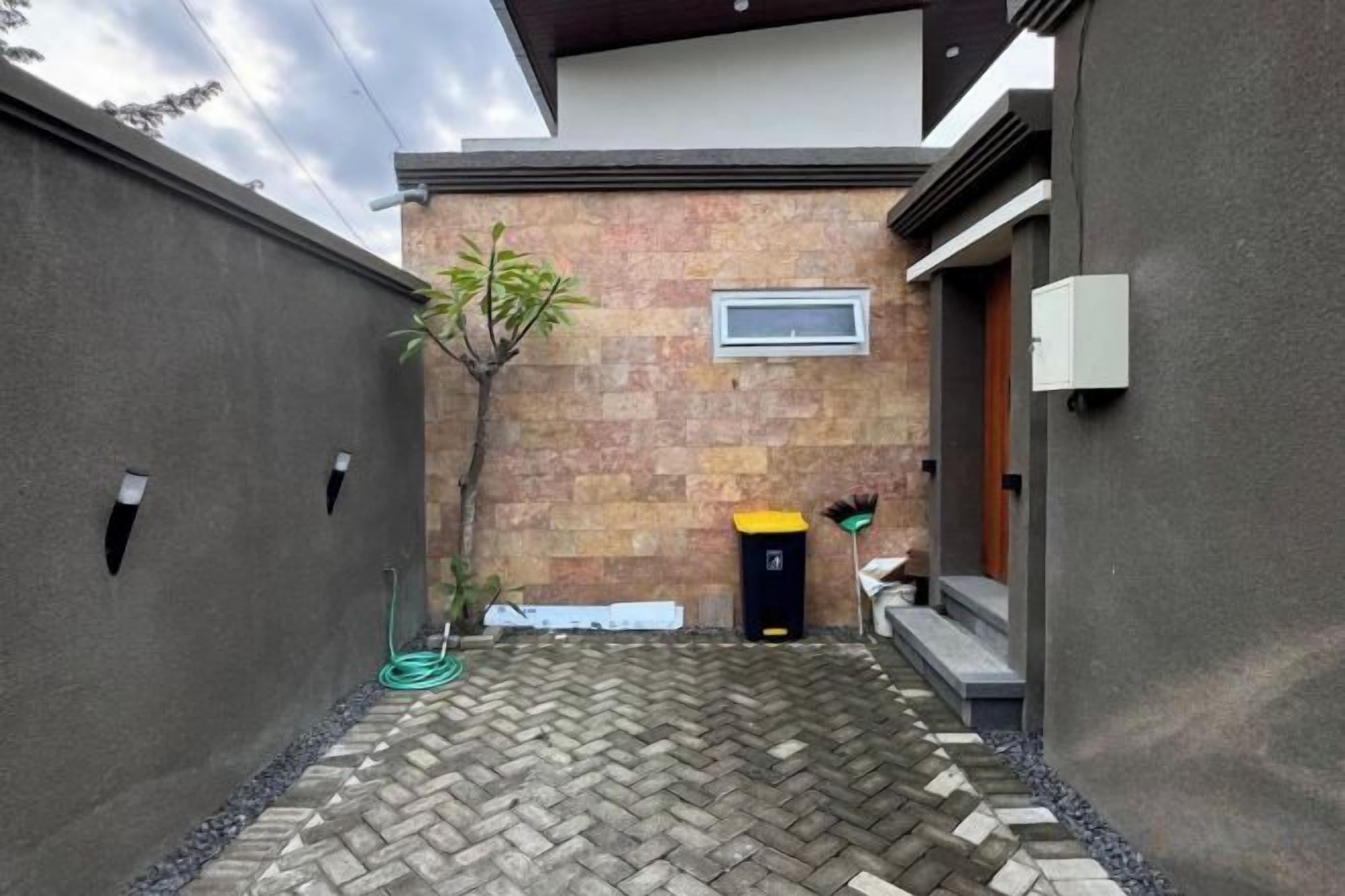 A narrow courtyard with a herringbone-patterned stone floor, a small tree, and a black trash bin.