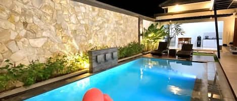 A swimming pool with a stone wall, a lounge area with chairs, and a well-lit outdoor space.