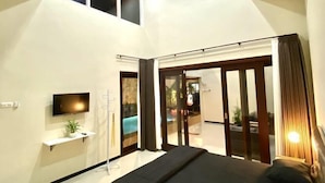 Une chambre moderne avec un lit, une télévision, une petite étagère ornée d’une plante et une porte-fenêtre coulissante donnant sur un balcon.
