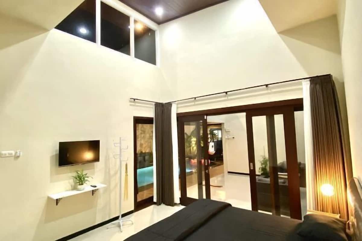 Een moderne slaapkamer met een bed, een televisie, een klein plankje met een plant en een glazen schuifdeur naar een balkon.
