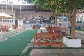 An outdoor seating area with wooden tables and chairs, a green turf, and a building with a sign in the background. - Azana BeSt Hotel Batu (Junrejo)