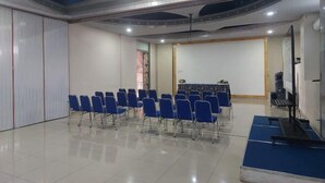A conference room with rows of blue chairs, a stage with a podium, and a large mirror on the wall. - Azana BeSt Hotel Batu (Junrejo)