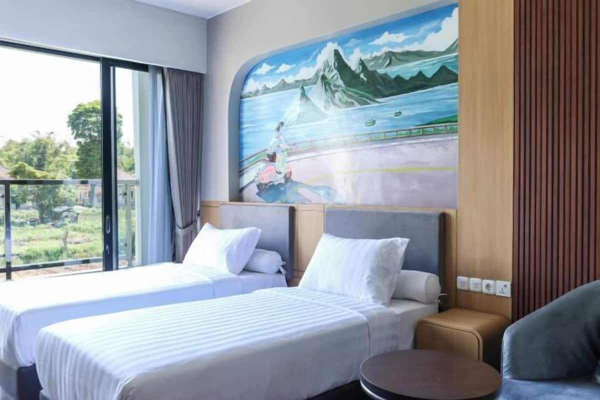 Habitación de hotel con dos camas, un sofá, una mesita y un mural de una persona en un patinete, además de un ventanal grande.