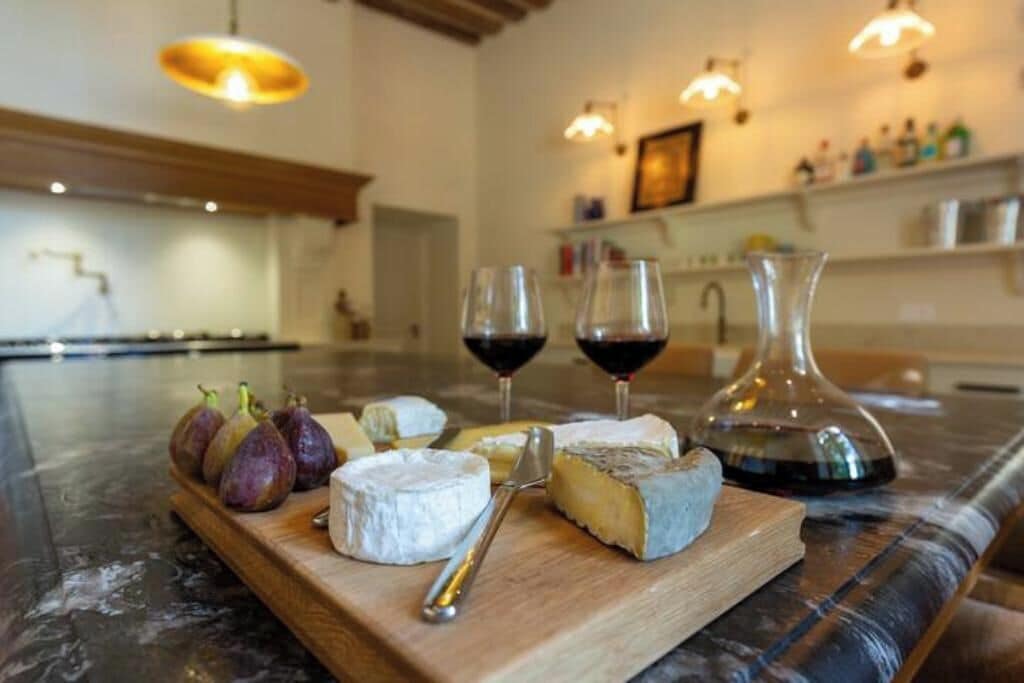 Tabla de quesos con diferentes tipos de queso e higos sobre una tabla de madera, acompañada de dos copas de vino tinto y una jarra de decantador sobre una encimera de mármol.
