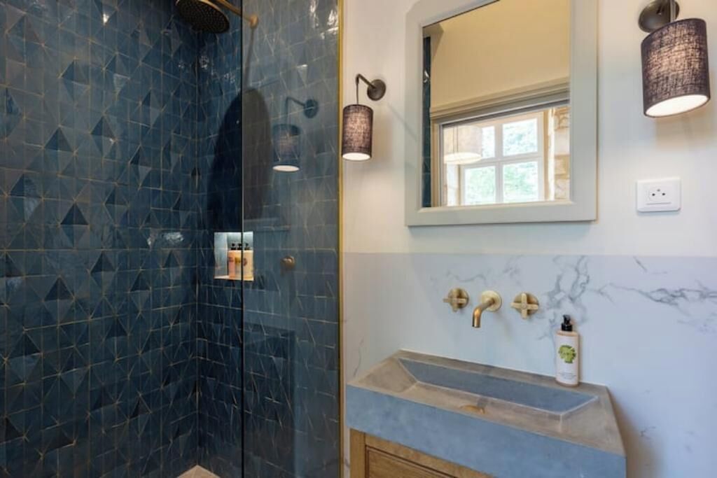 Une salle de bain avec une douche dont le carrelage est bleu, un lavabo en marbre et une fenêtre située au-dessus du miroir.