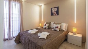A hotel room with a large bed, two bedside tables with lamps, a window with curtains, and a wall-mounted picture. - ONDA BIANCA LUXURY APARTMENTS (Realmonte)