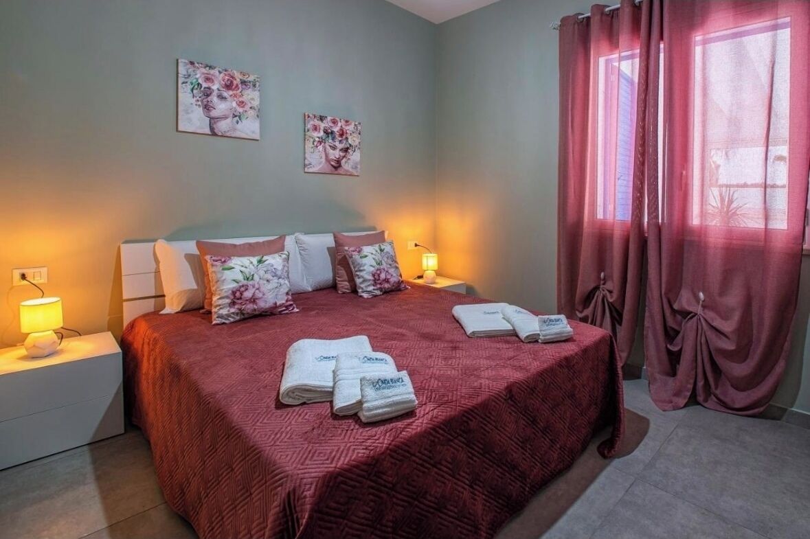 Un lit bien fait, recouvert d’une courtepointe rouge, deux tableaux accrochés au mur et une fenêtre ornée de rideaux roses.