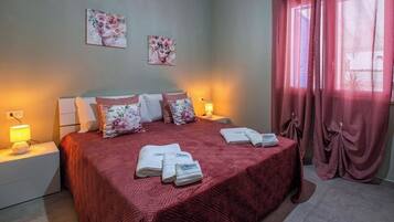 A neatly made bed with a red bedspread, two wall-mounted paintings, and a window with pink curtains.