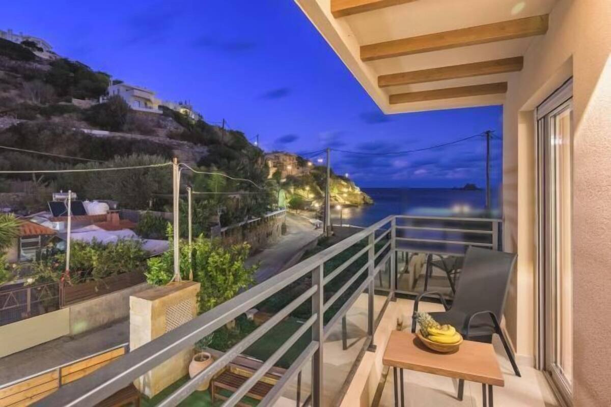 A balcony with a table and chairs, overlooking a coastal road and buildings.