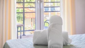 A bed with a rolled white towel on it, a window with a view of a patio and plants, and a door with a metal gate. - BlueGreen Holiday Homes: Holidays with Seaview (Malevizi)