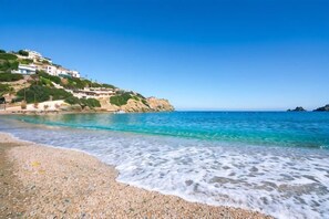 Una playa con aguas turquesas cristalinas, orilla de guijarros y edificios en una colina.