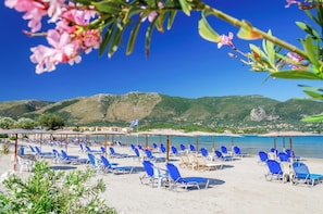 A beach with rows of blue sun loungers and umbrellas, a flag, and a mountainous backdrop.