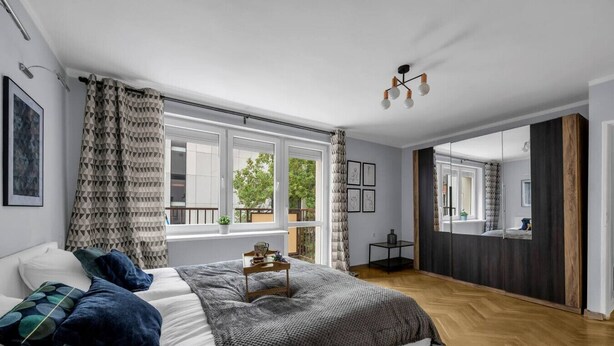 A modern bedroom with a large bed, a bedside table, a mirror, and a view of the outdoors through large windows.