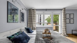 A modern bedroom with a large window, a bed with patterned pillows, a bedside table with a tray, and framed artwork on the walls.