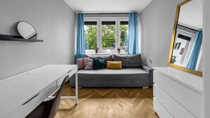 A modern, compact room with a grey sofa, a white desk, and a large window with blue curtains.