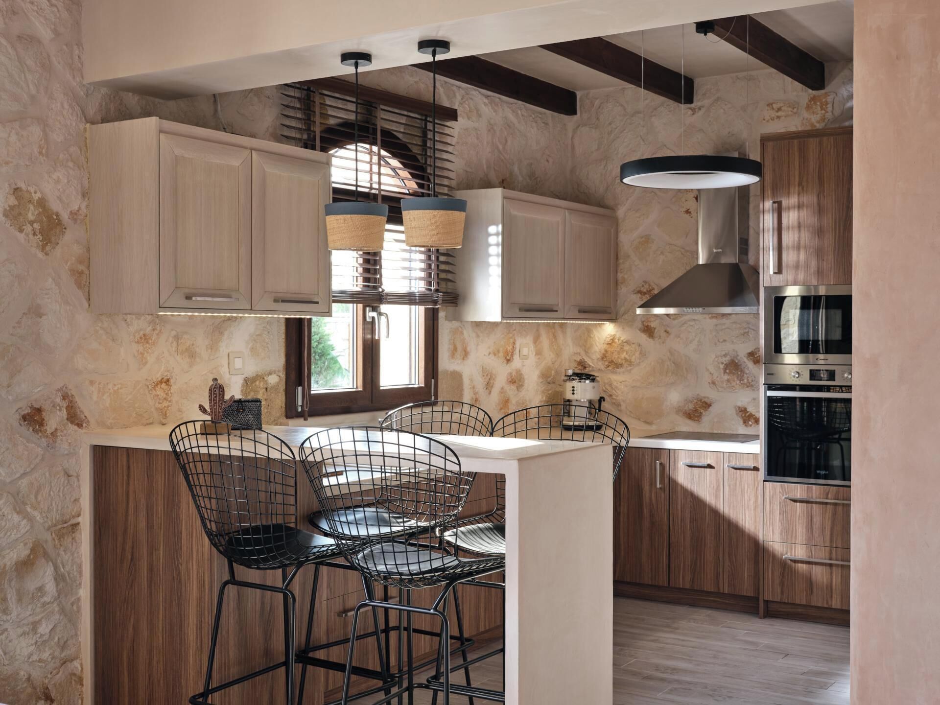 A modern kitchen with a stone wall backsplash, wooden cabinets, a built-in oven, and a bar area with metal chairs.