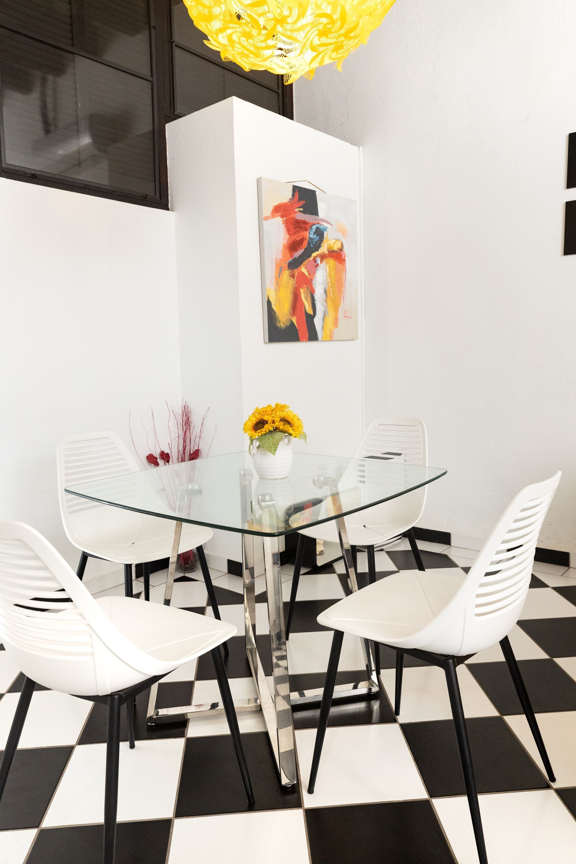 A modern dining area with a glass table, white chairs, a vase with sunflowers, and a colorful abstract painting on the wall.