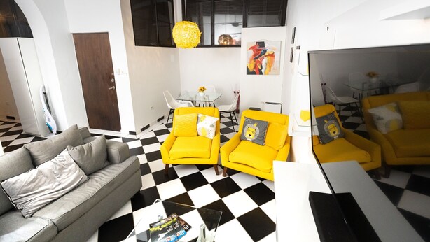 Een woonkamer met een zwart-wit geruite vloer, een grijze bank en twee gele fauteuils.