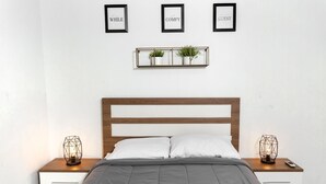 Una cama bien tendida con edredón gris, cabecera de madera y dos mesitas de noche con lámparas geométricas. En la pared, sobre la cama, hay tres letreros enmarcados.