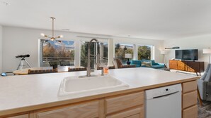 A modern kitchen with a large island, built-in dishwasher, and a view of mountains through large windows. - Modern Park Point - Walk to Town (Chelan)