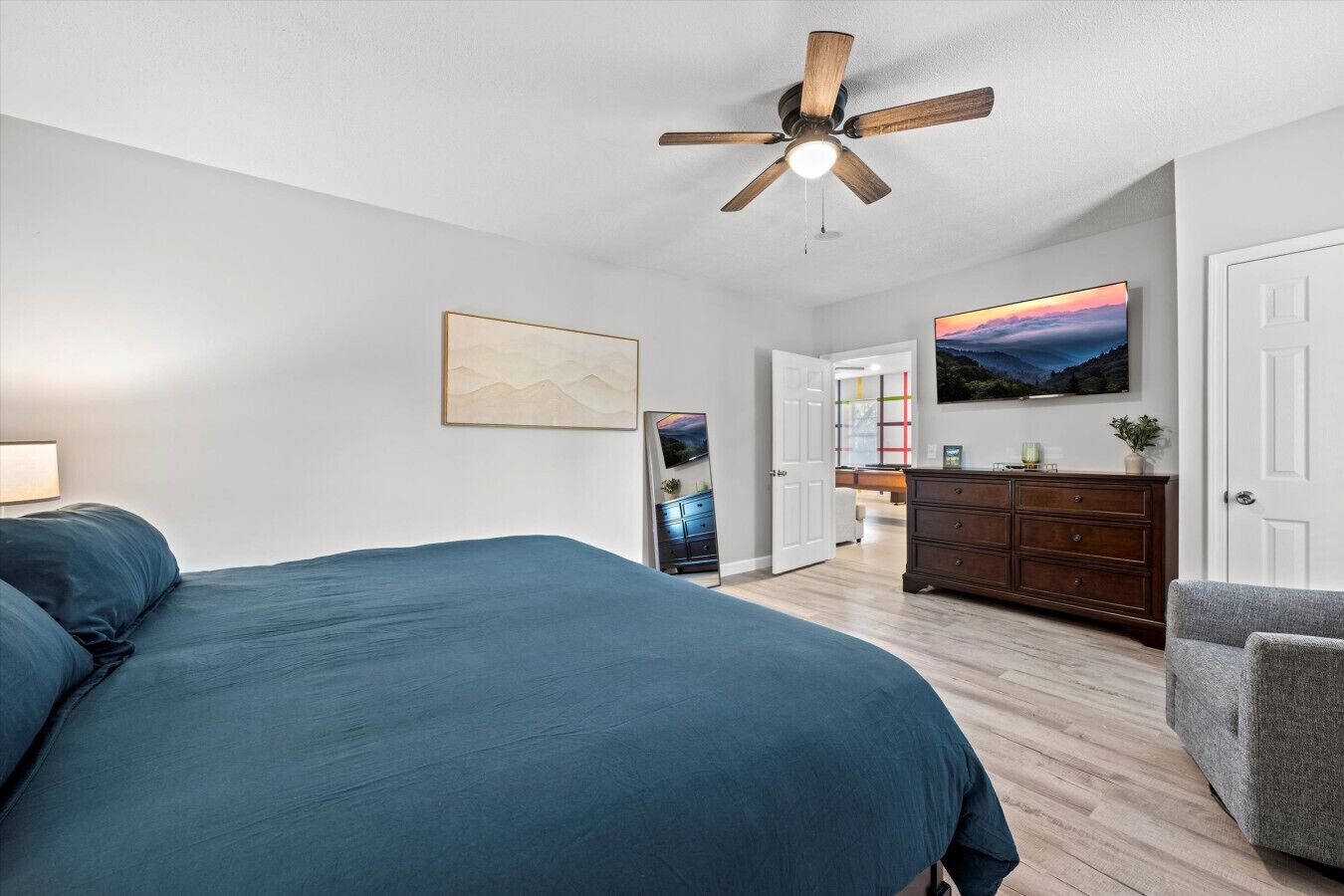 A bedroom with a large bed, a ceiling fan, a dresser, and a TV in the background.