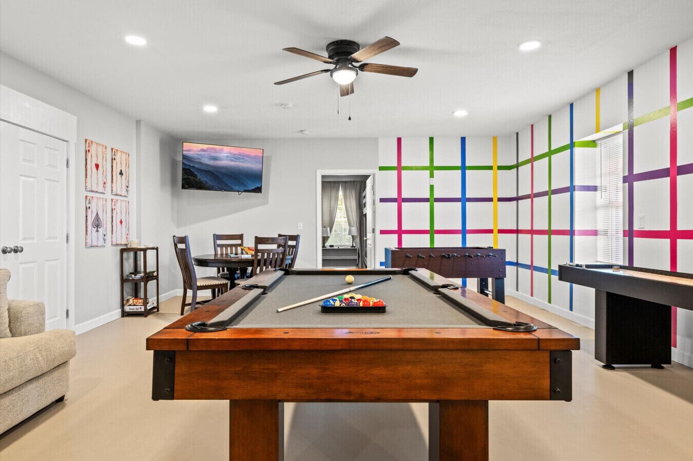 Una mesa de billar en una habitación con ventilador de techo, televisor y pared de colores.